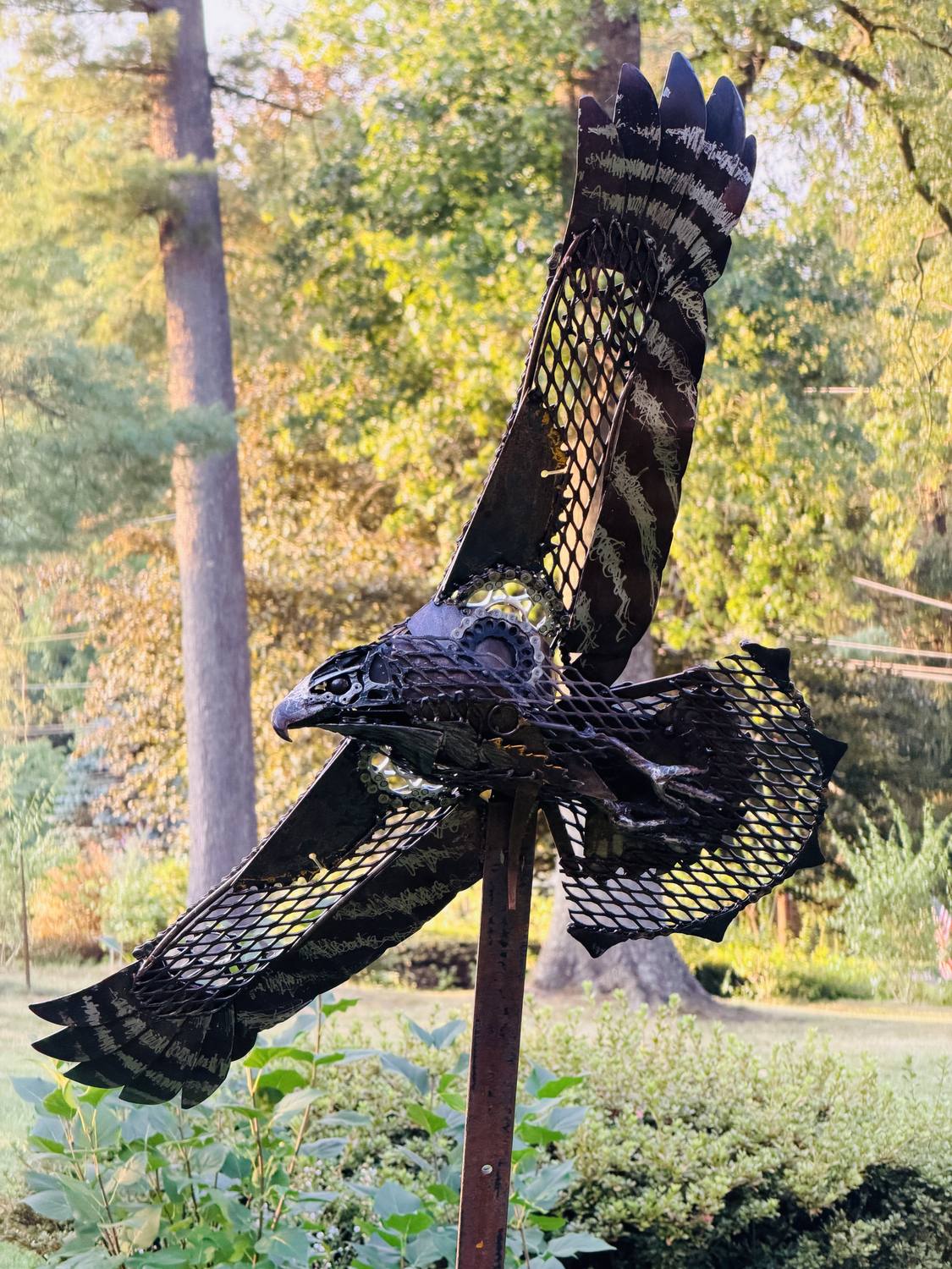Sentinel, a welded hawk sculpture with wings raised, mounted in a wooded garden